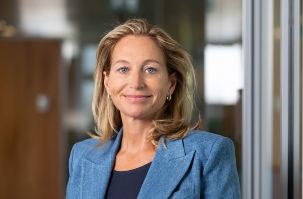 Susi in a blue blazer stands in a modern office setting with glass walls.