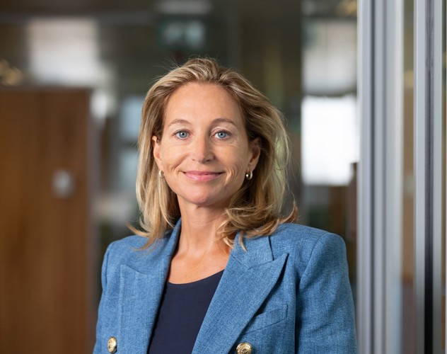 Susi in een blauwe blazer staat in een moderne kantoorruimte met glazen wanden.