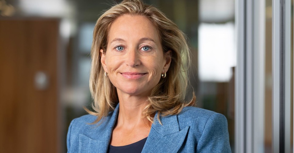 Susi in een blauwe blazer staat in een moderne kantoorruimte met glazen wanden.