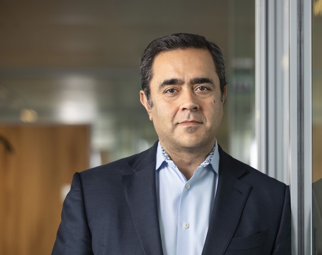 Qasim in een donker blazer en lichtblauw overhemd staat naast een glazen wand in een moderne kantooromgeving.