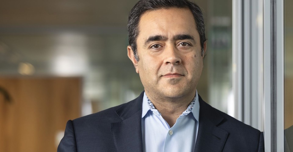 Qasim in een donker blazer en lichtblauw overhemd staat naast een glazen wand in een moderne kantooromgeving.