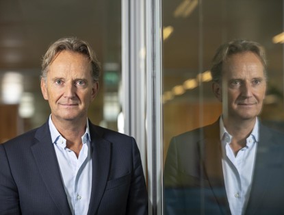 Joop in een donkerblauwe blazer staat voor een glazen wand met een reflectie in het glas.