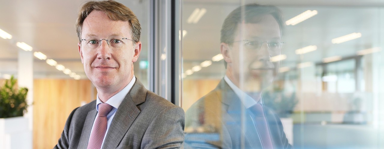 Reinout in a gray suit and a light pink tie stands next to a glass wall in a modern office setting.