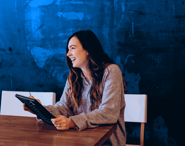 A woman in a gray sweater sits at a wooden table, looking at a tablet, with a blue wall in the background.
