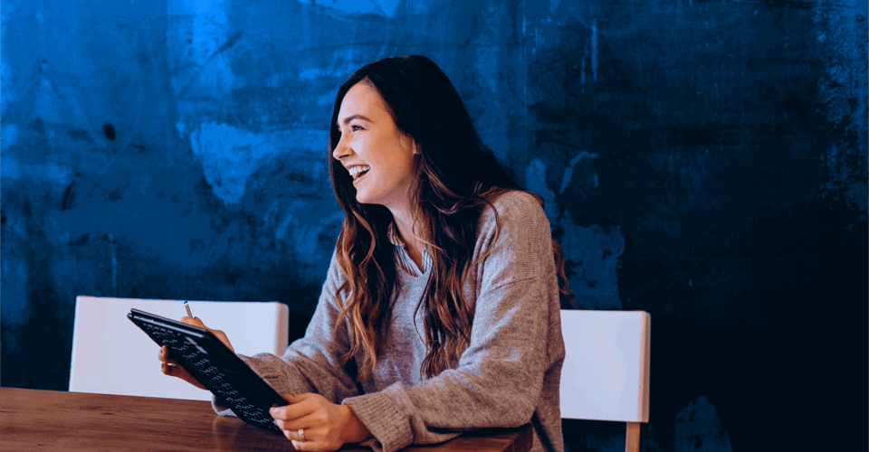 A woman in a gray sweater sits at a wooden table, looking at a tablet, with a blue wall in the background.