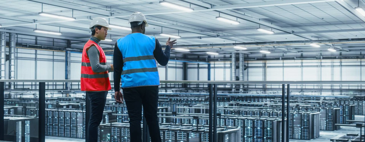 Twee mannen in veiligheidskleding, één met een rood en de ander met een blauw vest, kijken naar een datacenter vol servers.