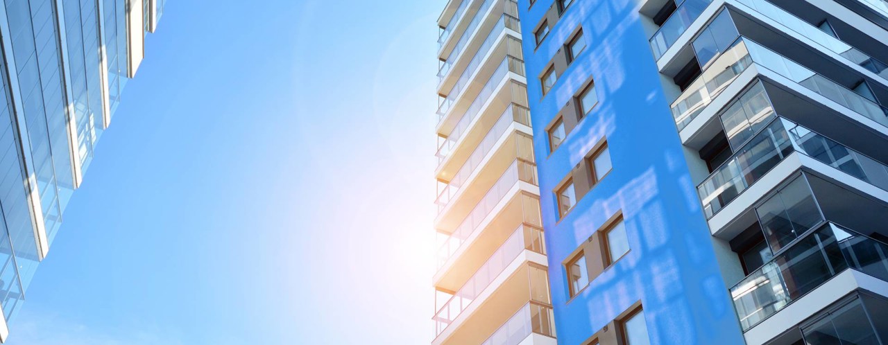 Hoog gebouw met glazen balkons en een blauwe gevel, tegen een heldere blauwe lucht met zon.