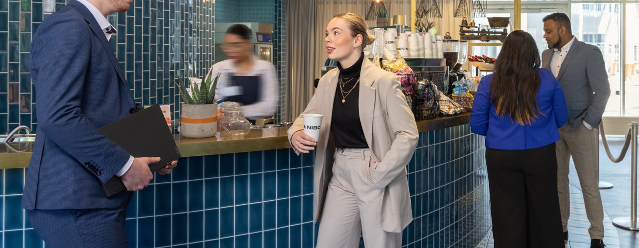 Een vrouw in een beige pak houdt een koffiebeker vast en praat met een man in een blauw shirt bij een moderne cafébar.
