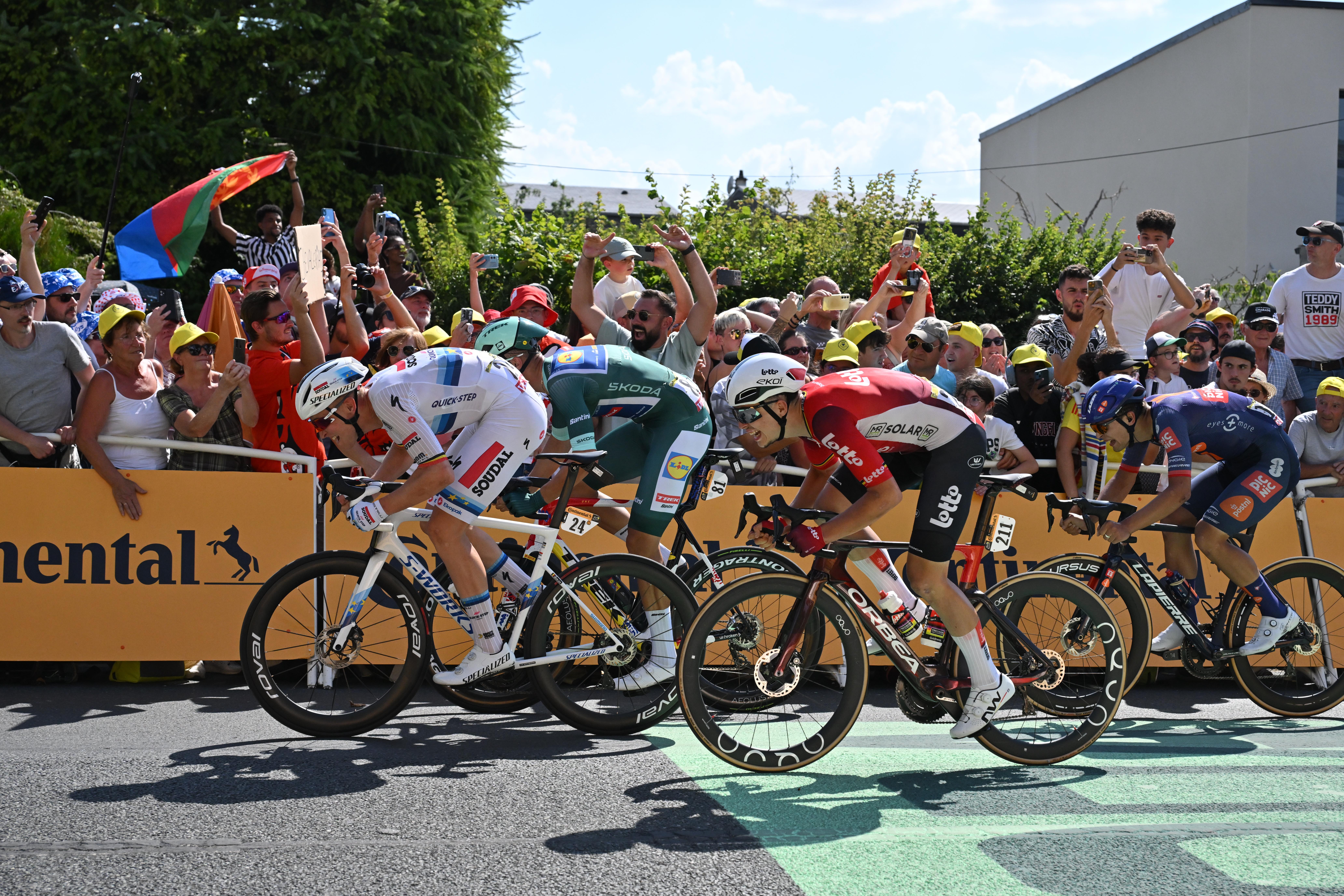 Road cyclists compete side by side during a race, surrounded by cheering fans holding flags and signs.