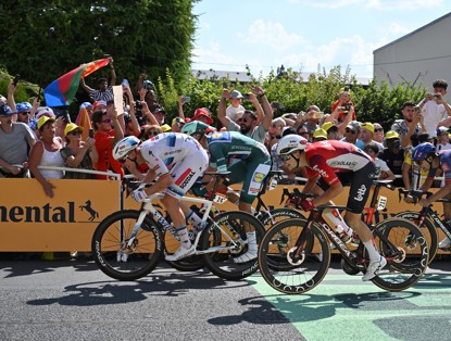 Road cyclists compete side by side during a race, surrounded by cheering fans holding flags and signs.