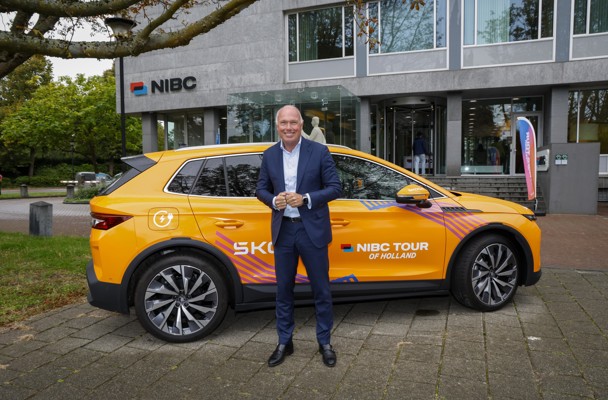 A man stands next to a brightly colored car displaying the NIBC Tour of Holland livery, in front of a modern office building.