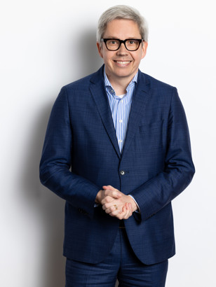 Toine in een blauw pak met gekruiste handen, staand voor een neutrale witte achtergrond.