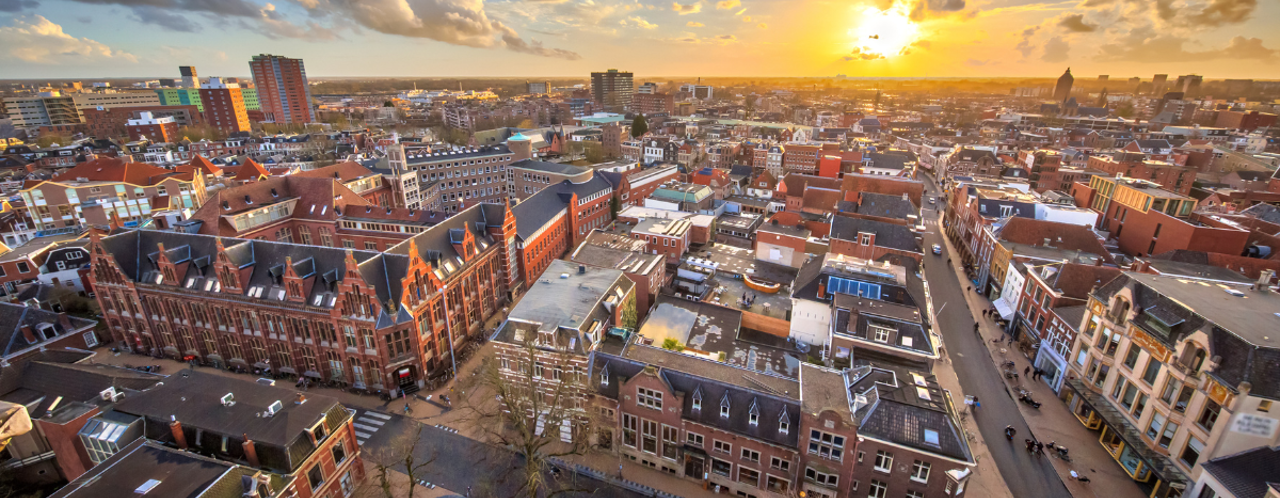 Uitzicht op een stad bij zonsondergang met historische gebouwen en moderne architectuur, onder een kleurrijke lucht.