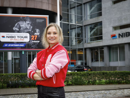 A person in a red varsity jacket poses in front of a large billboard for the NIBC Tour of Holland, visible in the background.